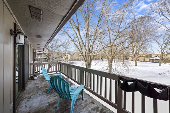 A balcony with blue chairs overlooks a snowy landscape with trees and neighboring houses.