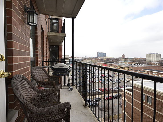 A balcony with chairs and grill overlooking a cityscape with buildings and parking areas.