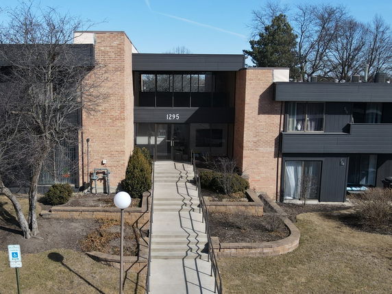 Front view of a building with brick and dark paneling, featuring a central entrance and landscaped walkway.