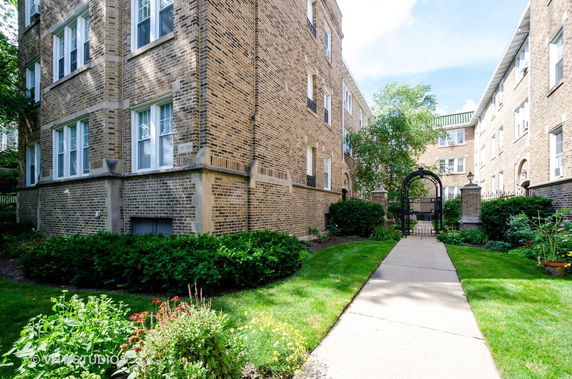 Front view of a brick apartment building with a garden and pathway leading to a gated entrance.