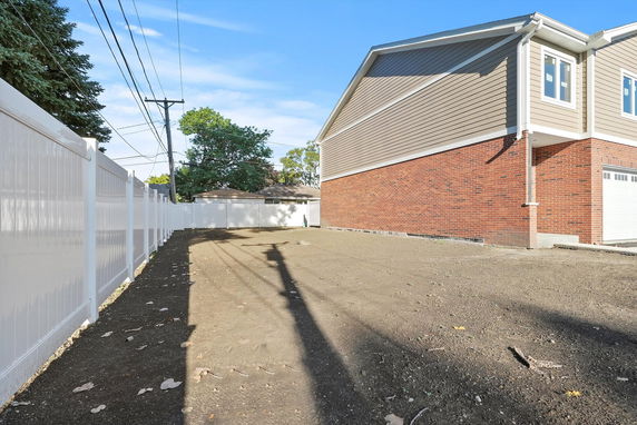 Side view of a two-story house with brick and siding exterior, adjacent to a dirt-covered area and white fencing.