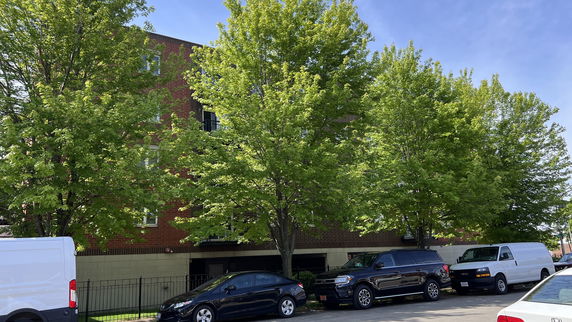 Front view of a multi-story brick building with trees and parked vehicles in front.