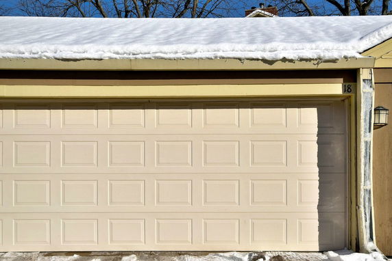 Front view of a garage with a closed light beige door and snow on the roof.