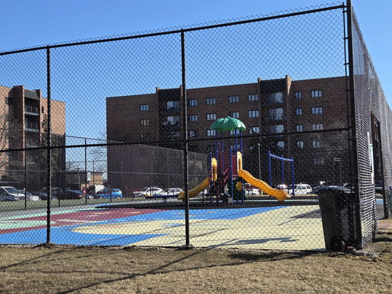 Playground with slides and climbing structures surrounded by a fence, with apartment buildings in the background.