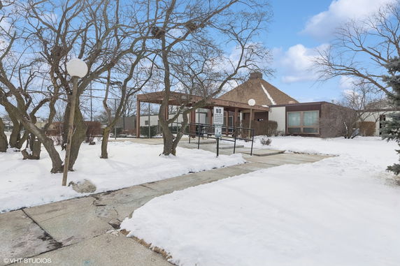 Front view of a building with a sloped roof and wood paneling, surrounded by trees and snow.