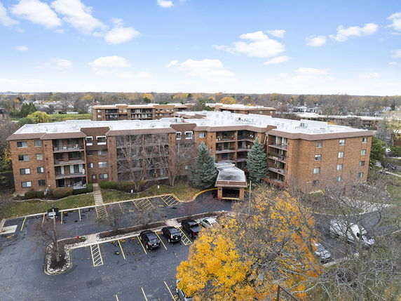 Aerial view of a multi-story brick apartment complex with parking area, surrounded by trees and other buildings.