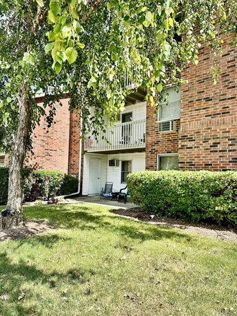 Front view of a multi-story brick apartment building with a small balcony and a grassy area with a tree.