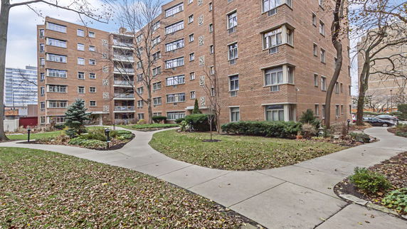 Front view of a multi-story brick apartment building with paths and gardens.
