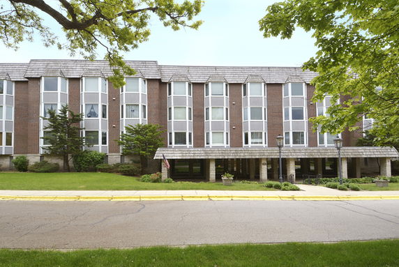 Front view of a three-story building with large windows and an entrance covered by an extended roof supported by columns.