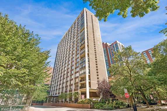 Front view of a multi-story residential building with balconies and a surrounding area with trees.