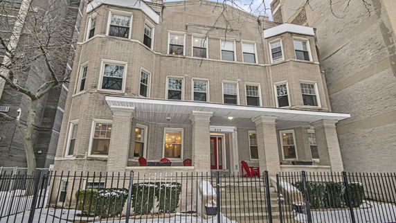 Front view of a three-story brick building with a covered porch and red chairs.
