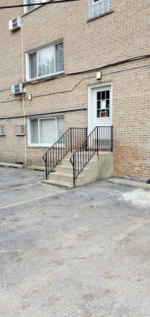 Exterior view of a building with brick facade, featuring steps leading to a door and adjacent parking area.