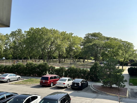 View of an outdoor area with parking and a fenced playground with trees in the background.