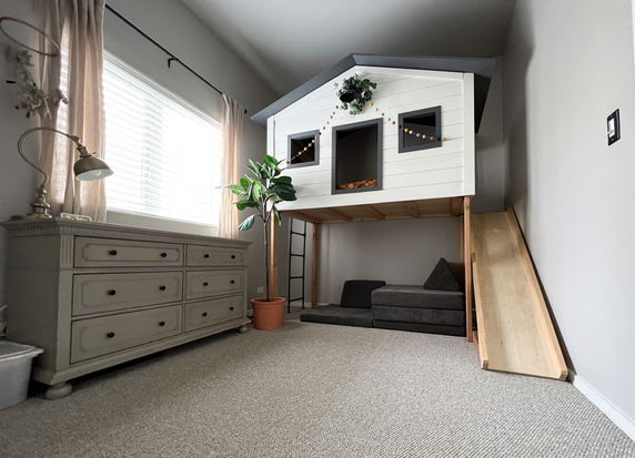 Indoor play area featuring a children's playhouse with a slide and a ladder inside a room with carpet flooring.