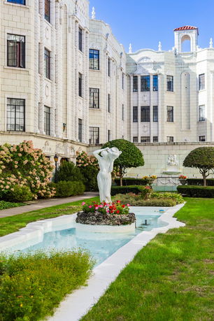 Front view of a multi-story building with ornate architectural details and a courtyard featuring a statue and fountain.