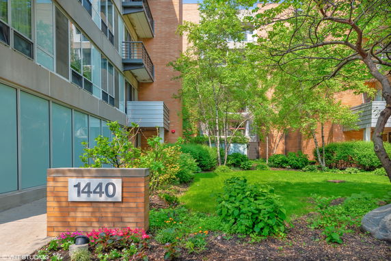 Front view of a multi-story building with a brick exterior and balconies, featuring a small landscaped area with greenery and a sign displaying the number 1440.