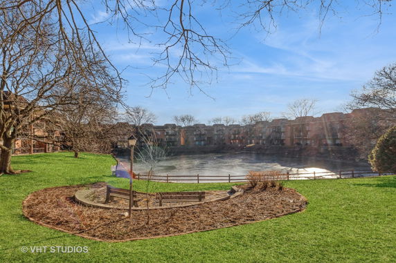 View of residential buildings surrounding a pond with trees and grass in the foreground.