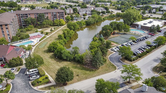 Wide aerial view of an apartment complex with surrounding greenery, a lake, swimming pools, and tennis courts.