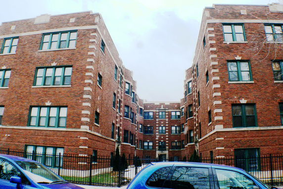 Front view of a brick apartment building with multiple windows and fenced entrance.
