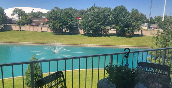 Panoramic view of a pond with a fountain, seen from a balcony with outdoor seating.