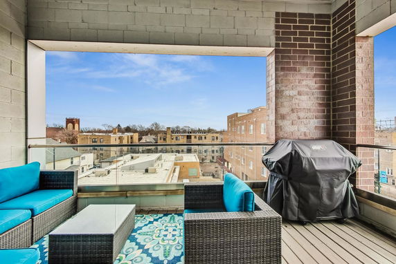 View from a balcony with outdoor seating and a barbecue grill, overlooking nearby buildings.