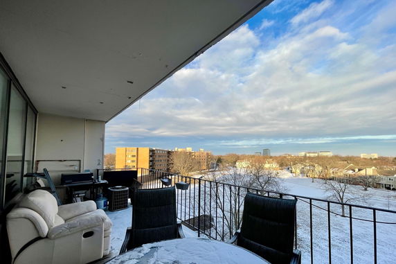 Snow-covered landscape viewed from a balcony with buildings and trees in the background.