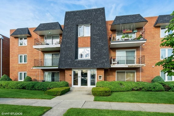 Front view of a three-story brick apartment building with balconies and a distinctive roof design.