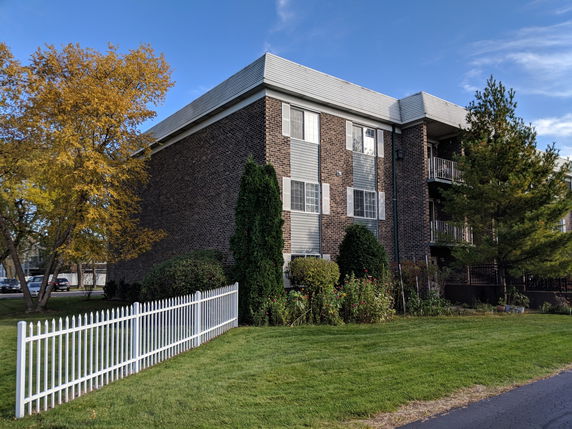 Front view of a three-story brick apartment building with a white fence and green lawn.