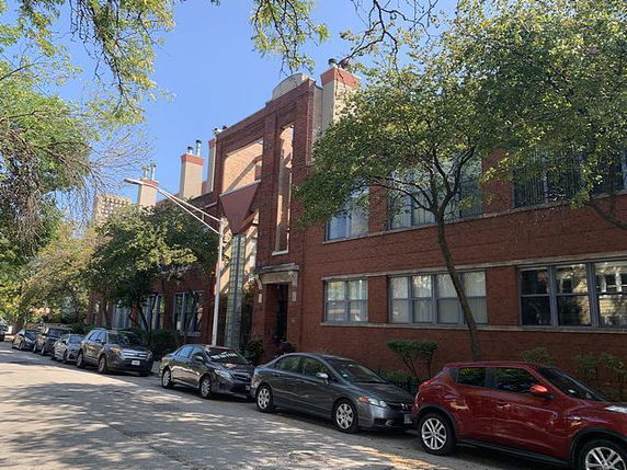 Front view of a multi-story brick building with trees and parked cars along the street.