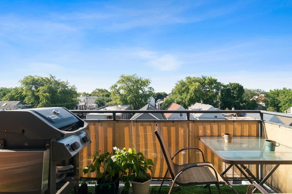 View from a balcony with a grill, chairs, table, and surrounding rooftops and trees.