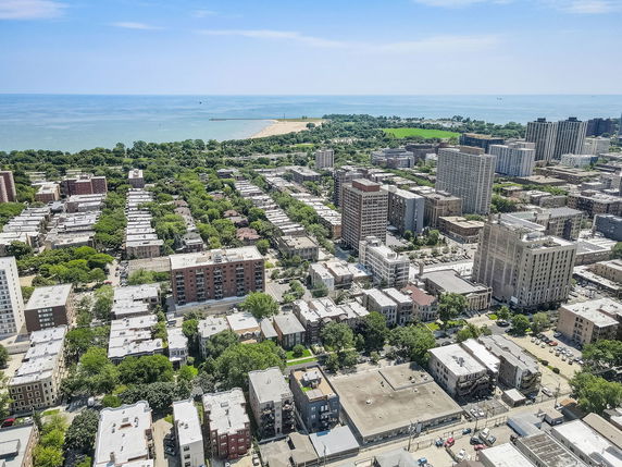 Aerial view of an urban area with a mix of residential and high-rise buildings, surrounded by greenery and overlooking a distant body of water.