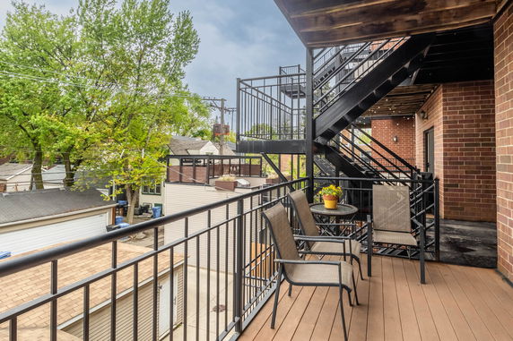 Outdoor view from a balcony with metal railings and outdoor seating, surrounded by brick walls and external stairs.