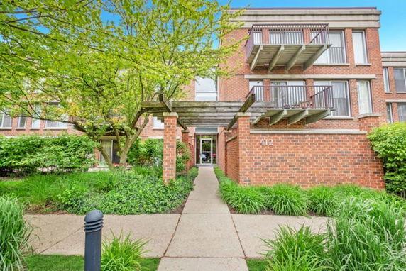 Front view of a brick apartment building with balconies and a walkway lined with greenery.