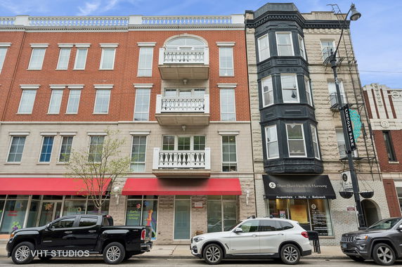 Front view of a multi-story building with red brick and white accents, featuring balconies and retail spaces on the ground level.