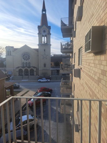 View from a building's balcony showing a street, parked cars, and a church with a tall spire.