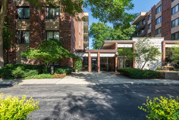 Front view of a brick apartment building with a glass entryway and surrounding greenery.