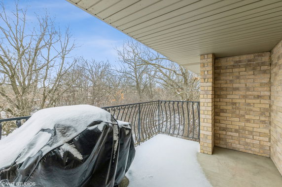 View from a covered balcony with a snow-covered grill and metal railing overlooking bare trees and a wintry landscape.