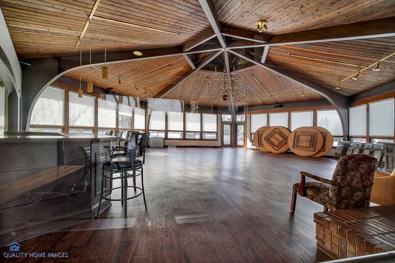 Interior view of a spacious room with a wooden ceiling and large windows, featuring chairs and tables stacked against the walls.