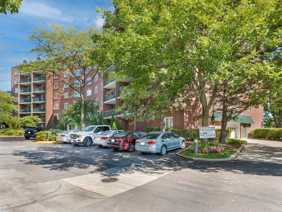 Outdoor parking area with multiple cars parked in front of a multi-story brick apartment building.