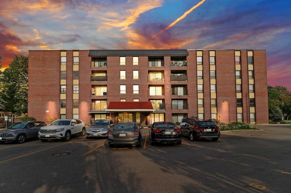 Front view of a multi-story brick apartment building with balconies and a parking area.