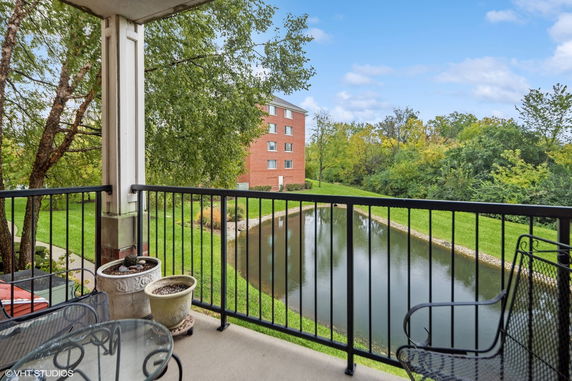 View of a pond and surrounding greenery from a balcony, with a view of a multi-story brick building in the background.
