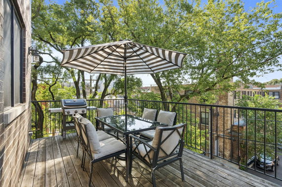 Outdoor deck with seating area and umbrella providing a view of trees and nearby buildings.