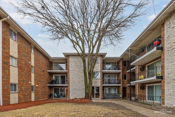 View of an apartment building with three stories, balconies, and a tree in front.