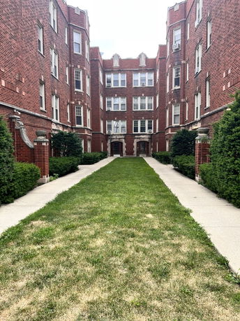 Front view of a multi-story brick apartment building with a grassy walkway leading to the entrance.