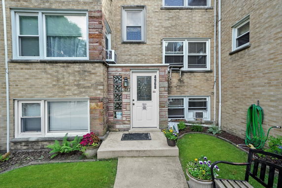 Front view of a multi-story brick apartment building entrance with potted plants and small lawn area.