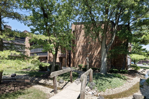 Front view of a multi-story brick building with trees and a small bridge in the foreground.