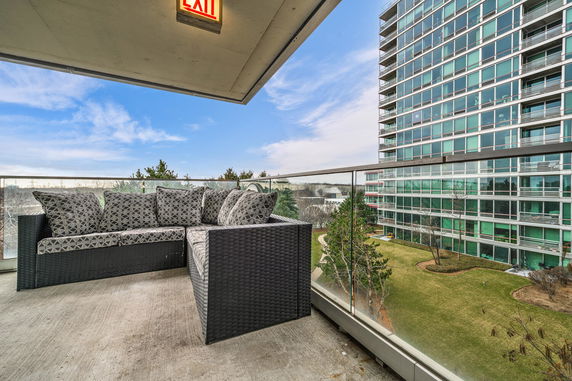 Balcony with a sofa and glass railing, overlooking a multi-story building and landscaped garden.