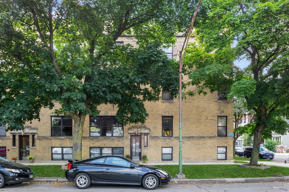 Front view of a multi-story brick building with parked cars and trees in front.