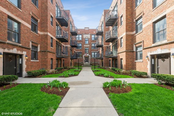 Front view of a multi-story brick apartment building with balconies and a landscaped courtyard.