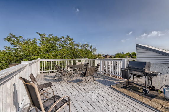 Rooftop with seating area and grill, surrounded by railing and trees.